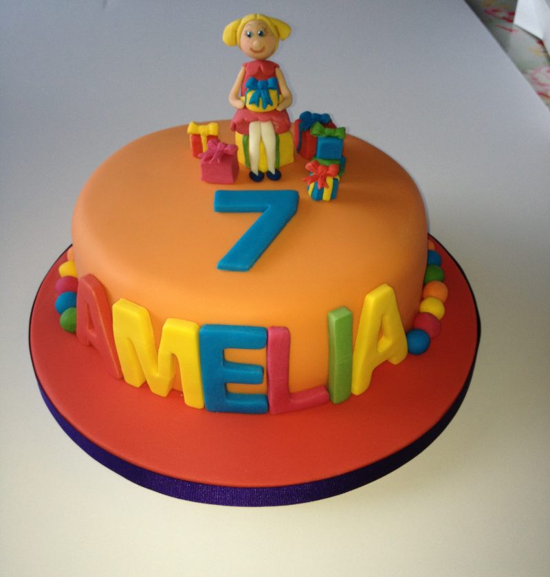 Bright birthday cake with girl sitting on the top and the name Amelia on the side